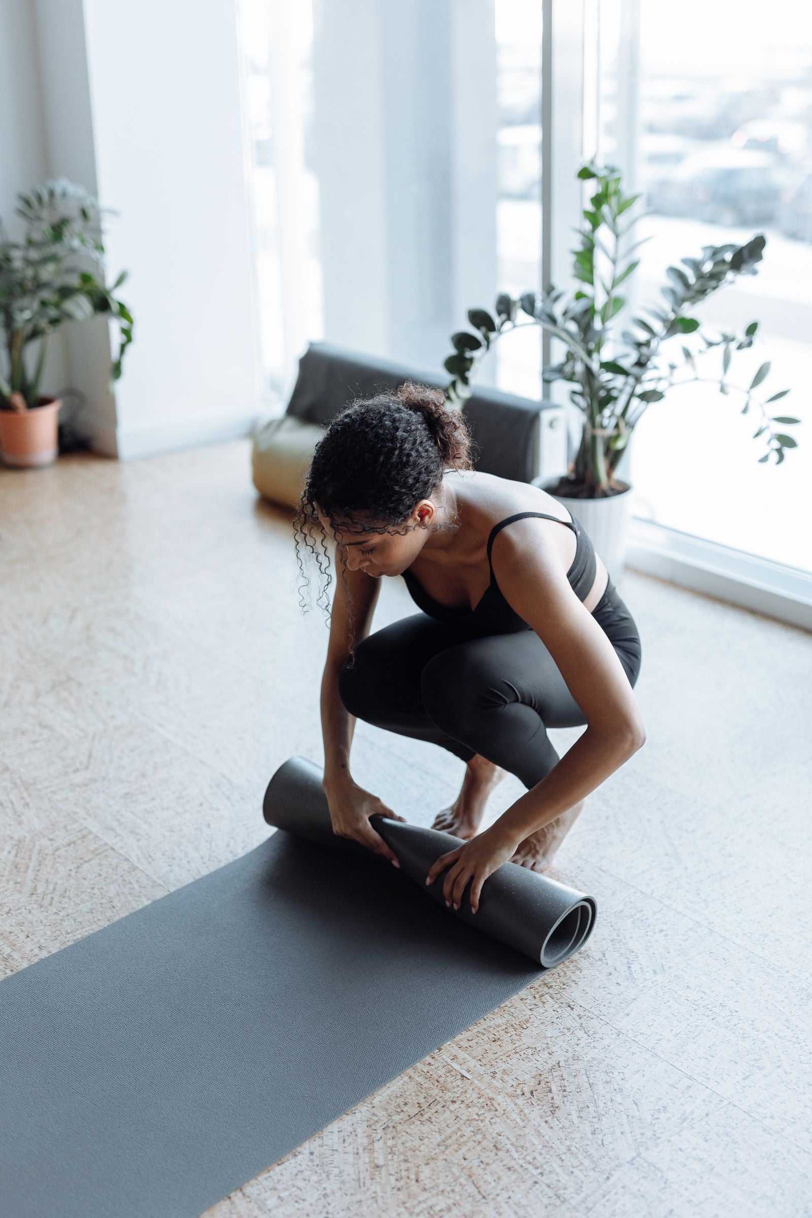 Woman rolling up a yoga mat in a sunlit studio surrounded by plants